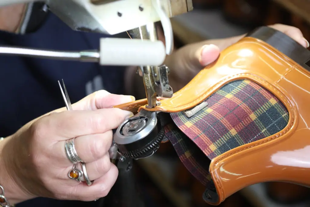 Mains d’une piqueuse guidant sous l’aiguille de la machine une botte de pluie Méduse couleur cognac à soufflet en tartan, dans l’atelier de Beaupréau