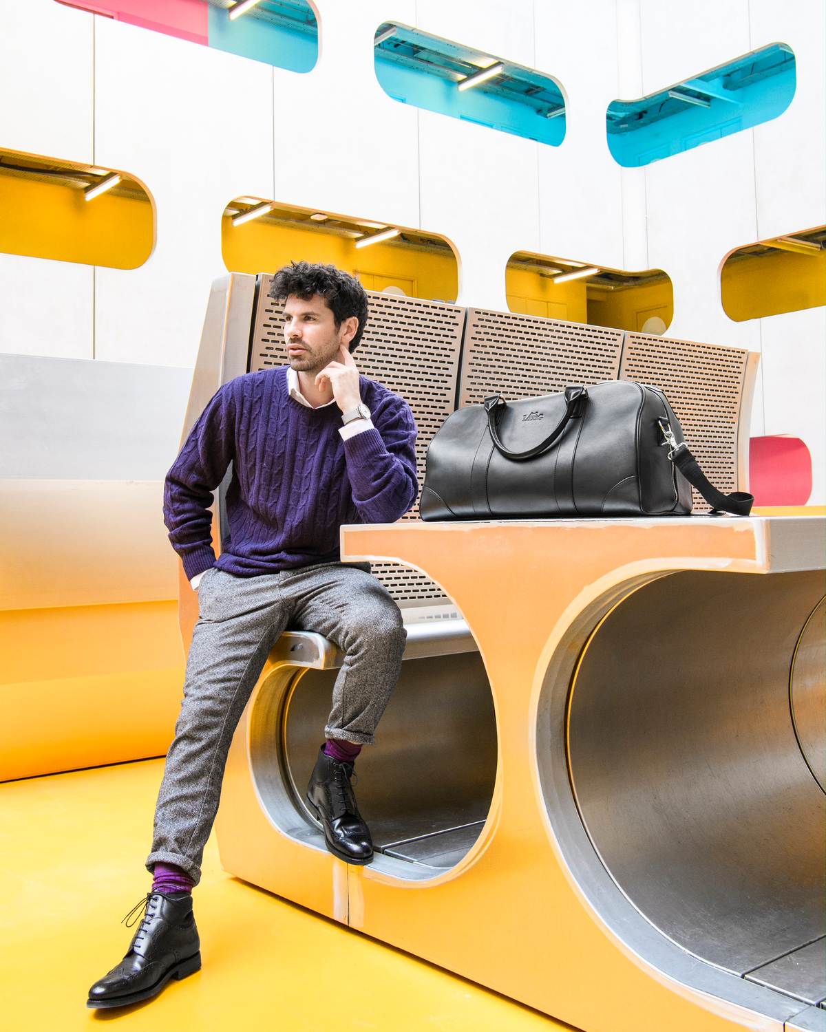 Un homme en pull violet assis dans un décor graphique jaune, un sac de voyage en cuir posé à côté de lui