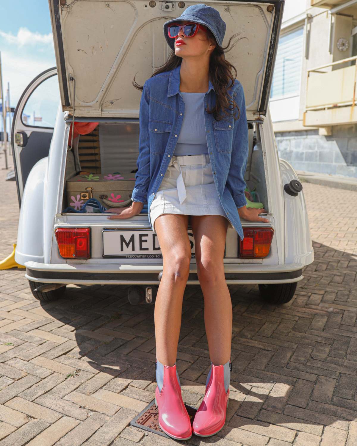 Une jeune femme en bob et veste en jean, assise dans le coffre d’une voiture ancienne, chaussée de bottines roses