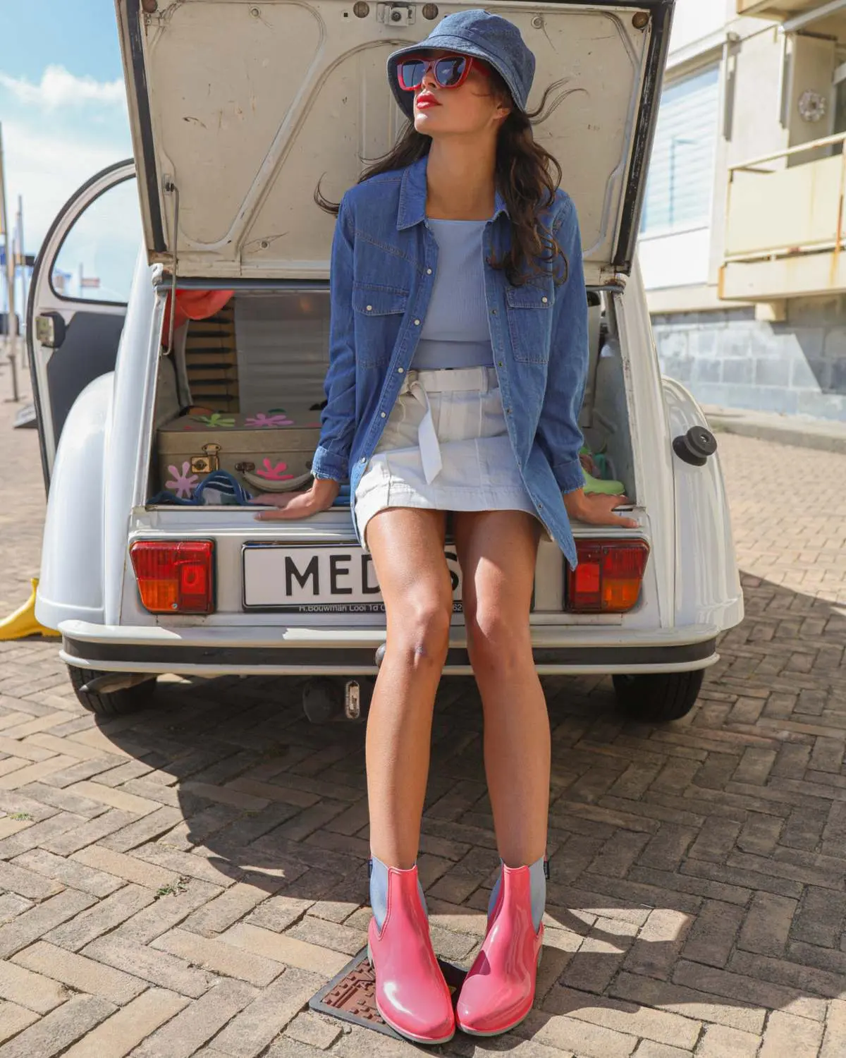 Une jeune femme en bob et veste en jean, assise dans le coffre d’une voiture ancienne, chaussée de bottines roses