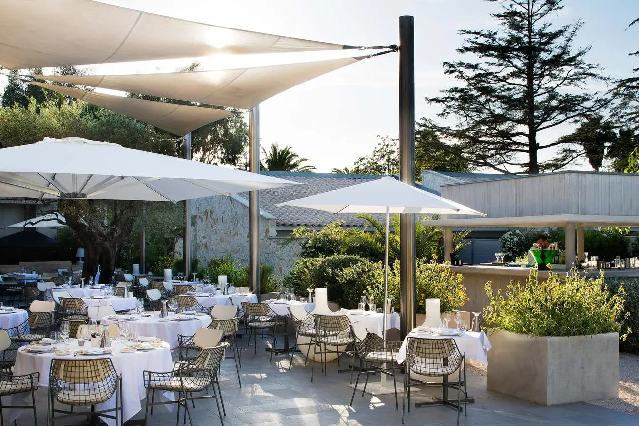 Terrasse du restaurant Colette au Sezz Saint-Tropez, tables nappées de blanc sous des voiles d’ombrage et des parasols, pins en fond