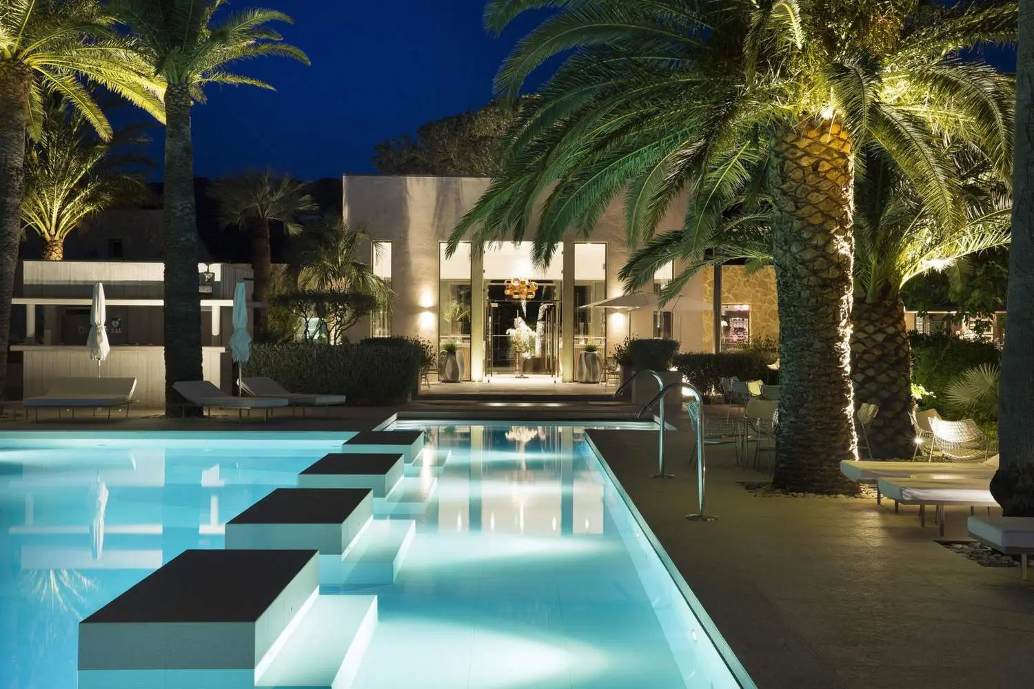 Piscine de l’hôtel Sezz Saint-Tropez de nuit, pas japonais éclairés sur l’eau turquoise, palmiers illuminés et entrée vitrée en fond