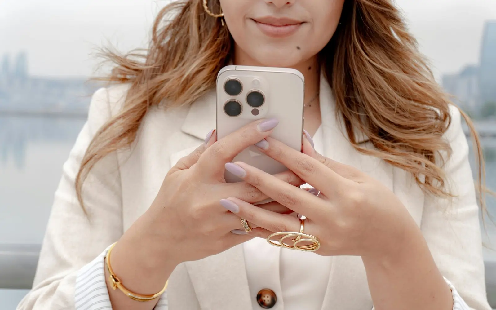 Une créatrice de contenu en veste crème tient son téléphone, mains ornées de bagues dorées