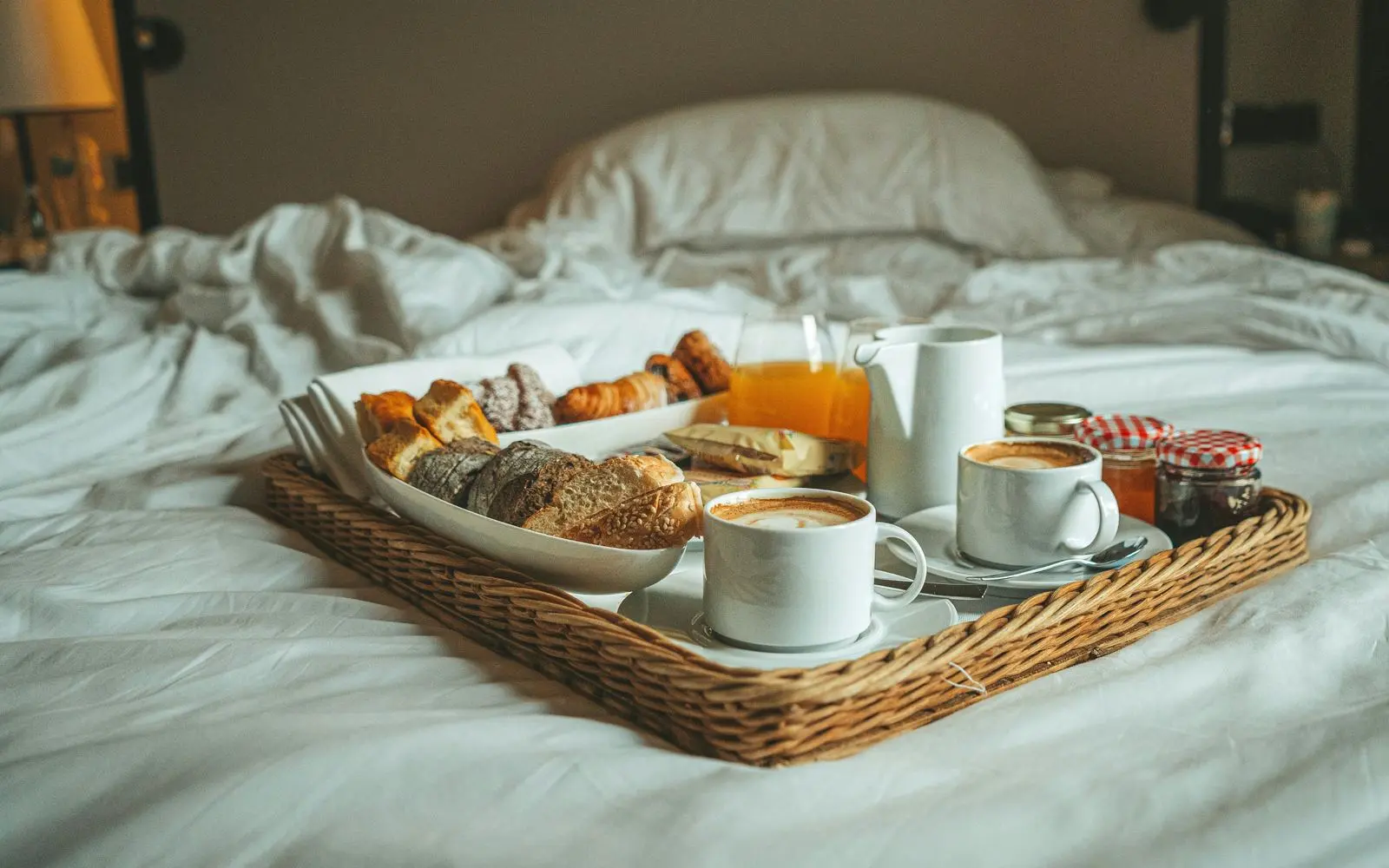 Un plateau de petit-déjeuner, viennoiseries, cafés et confitures, posé sur un lit aux draps blancs