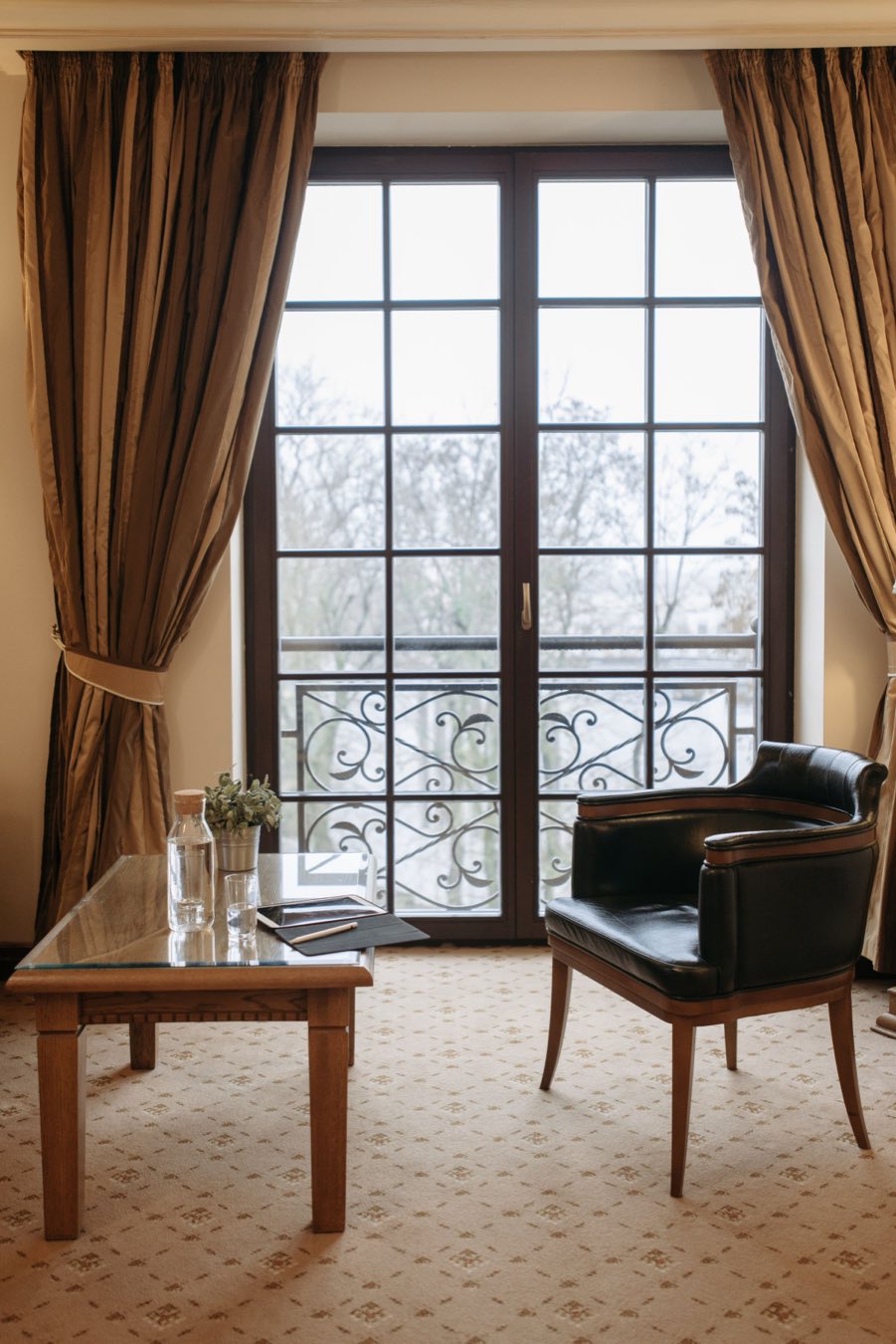 Fauteuil de cuir et table basse devant une porte-fenêtre à balcon ouvragé, encadrée de rideaux de soie